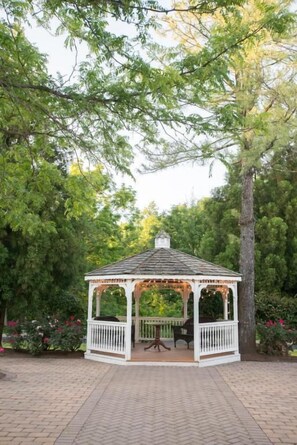 A gazebo with a table and chairs, surrounded by trees and a brick pathway. - Chester Hotel & Conference Center (Exton)