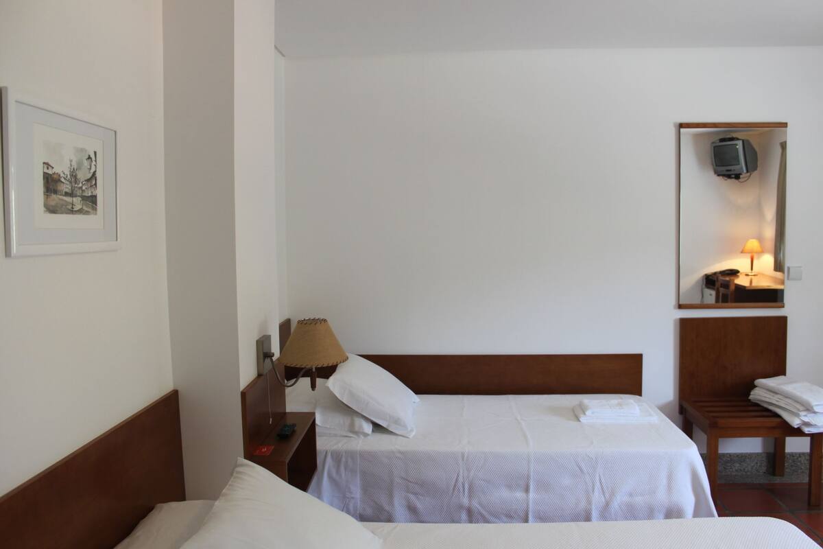 A hotel room with two beds, a bedside table with a lamp, a chair, a TV mounted on the wall, and a framed picture on the wall.