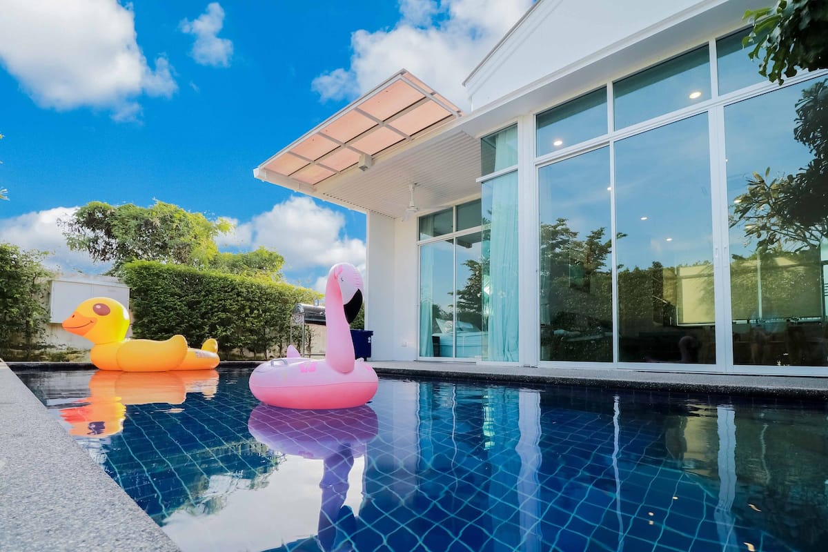 Una piscina con juguetes inflables, un edificio moderno con grandes ventanales y un cielo azul despejado.