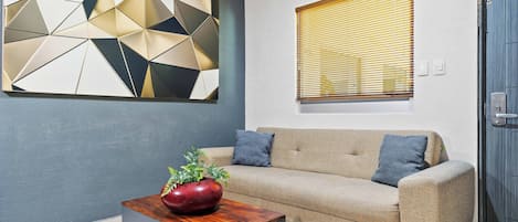 A modern living room with a beige sofa, a geometric wall art piece, a wooden coffee table with a plant, and a window with blinds.