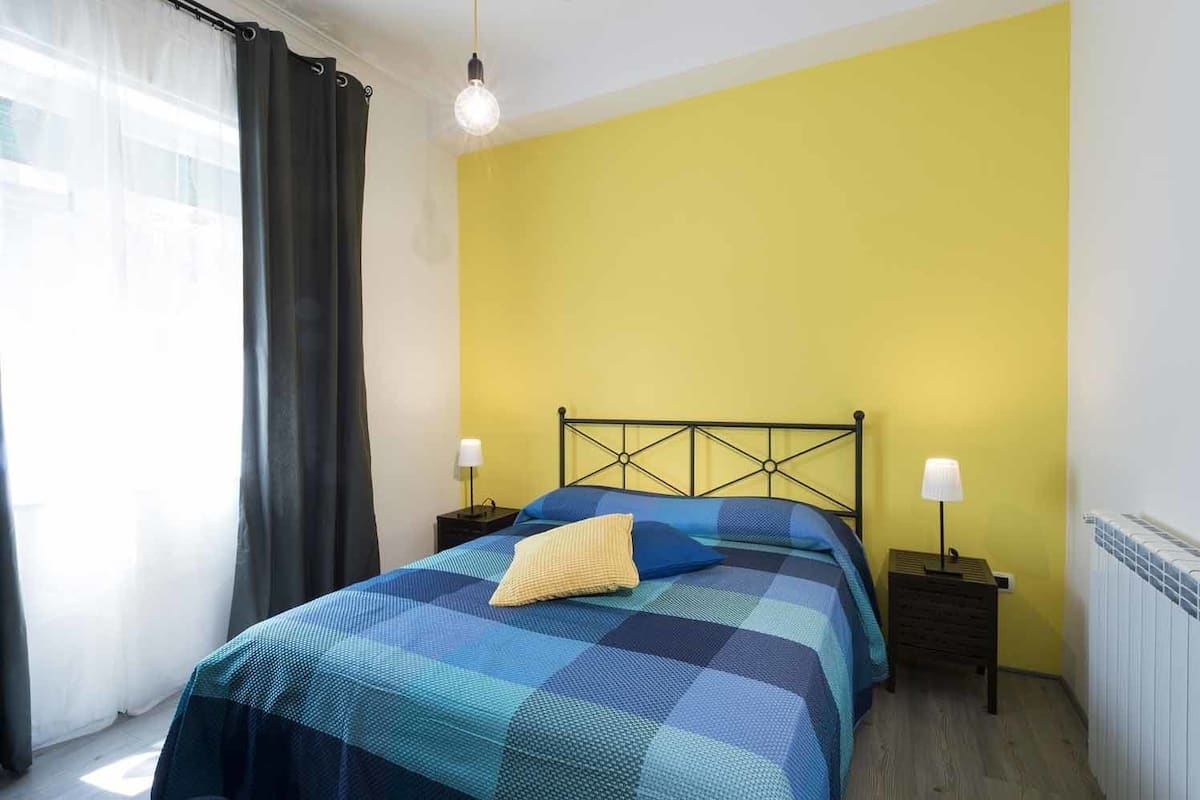 Un dormitori amb un llit, dues taules de nit, una finestra amb cortines i una paret groga.