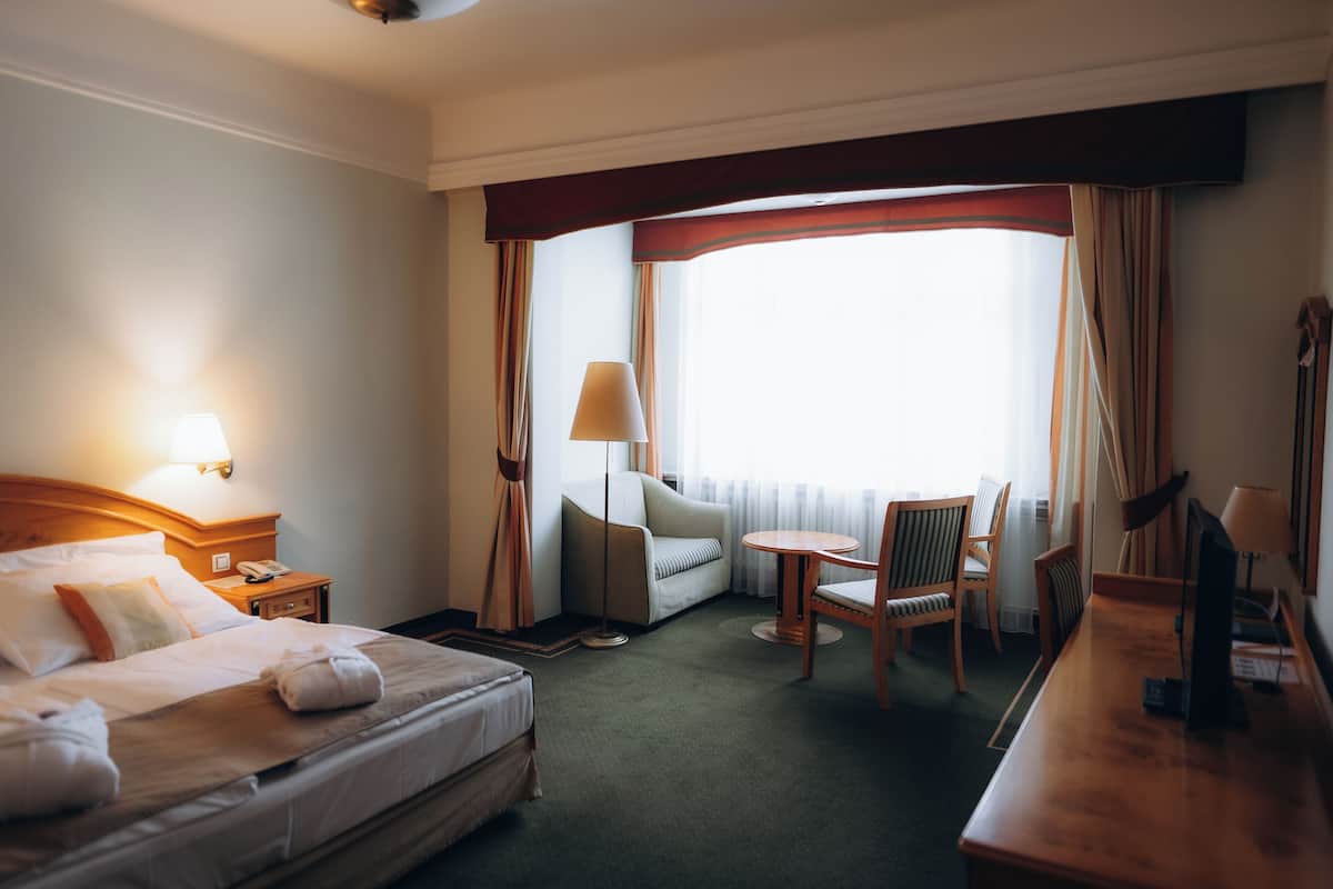 Een hotelkamer met een bed, een bureau, een stoel, een lamp en een raam met gordijnen.