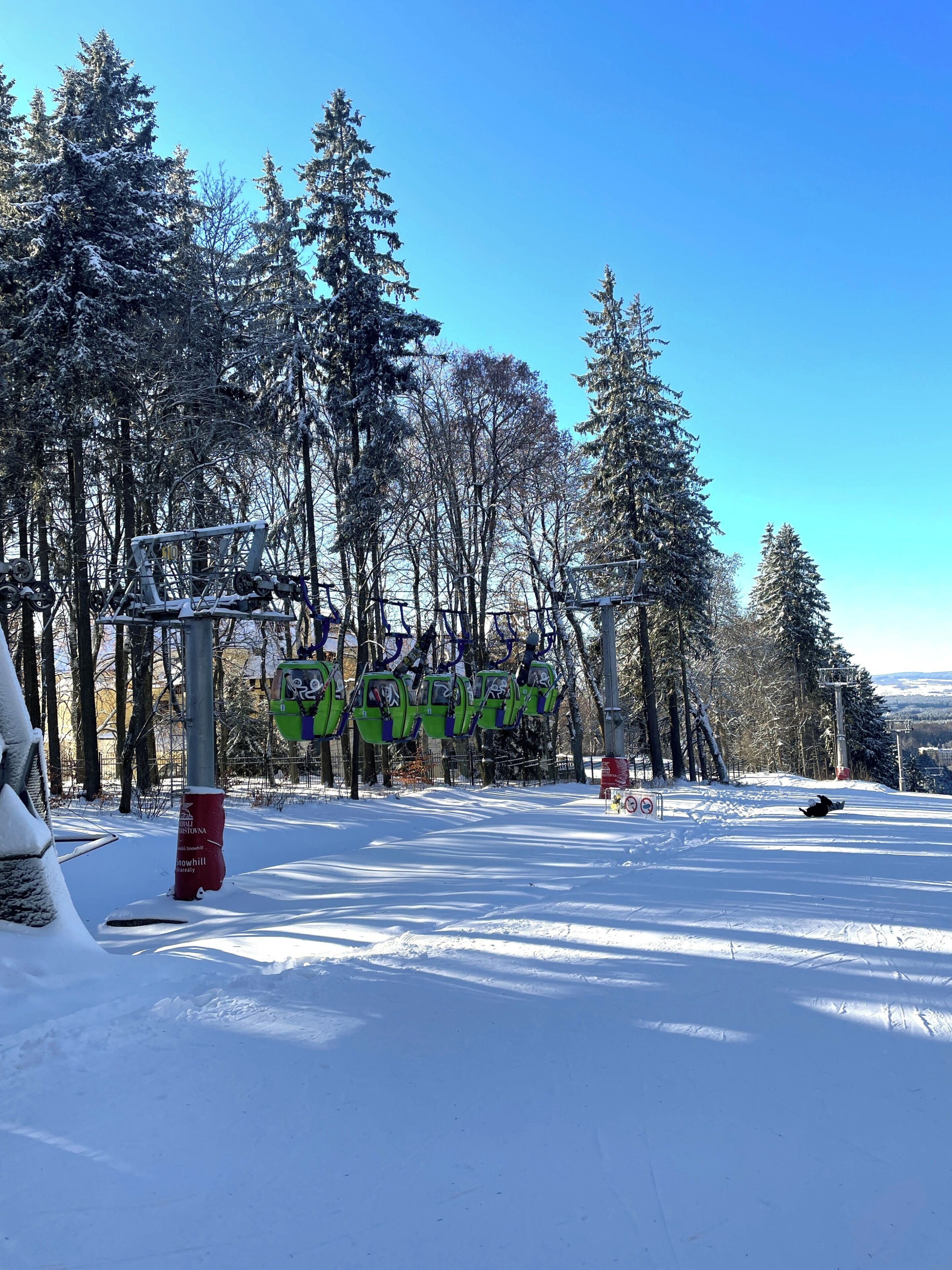 Estación de telesilla con sillas verdes, un cartel y árboles cubiertos de nieve.