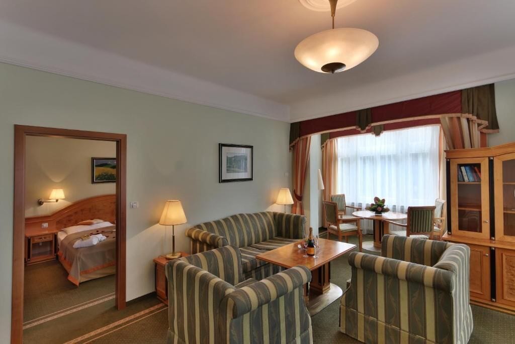 Une chambre d’hôtel comprenant un lit, un coin salon avec des fauteuils à rayures et une table, un meuble en bois et une fenêtre avec des rideaux.