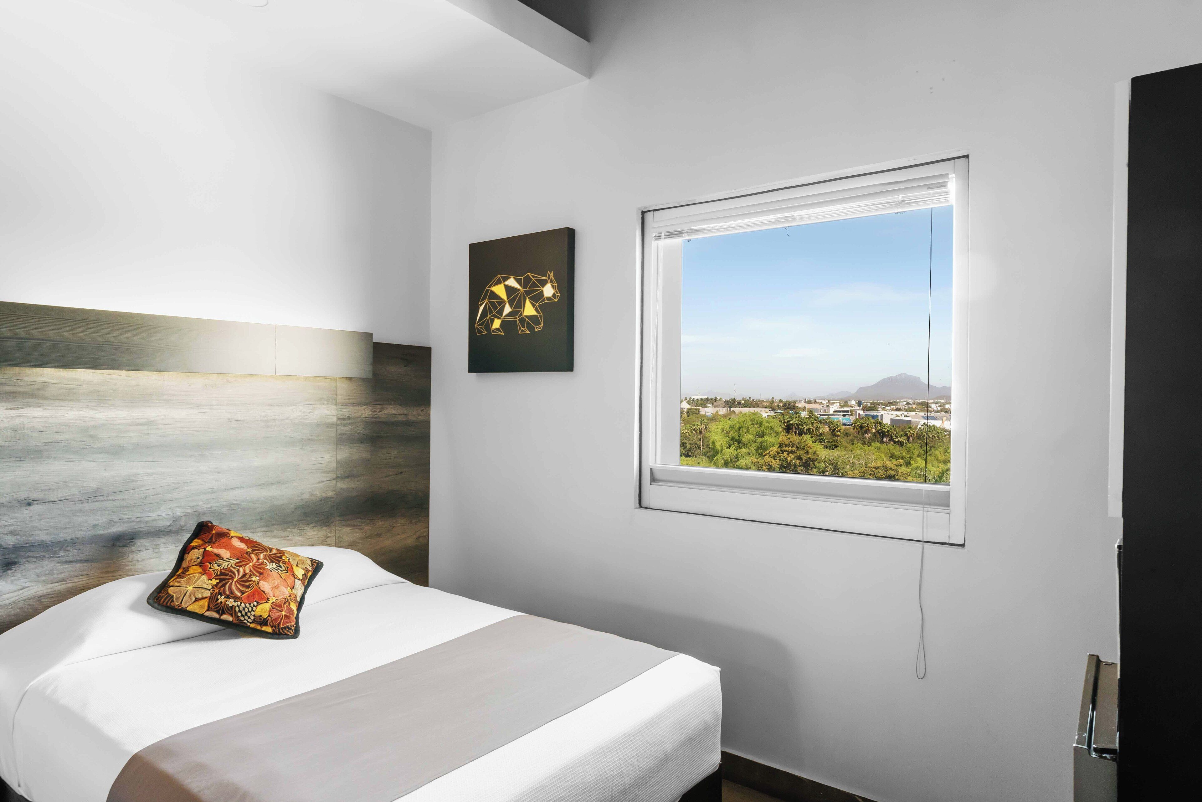 Una camera d'albergo moderna con un letto, vista sulla città attraverso una finestra e un'opera d'arte geometrica appesa al muro.