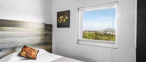 Habitación de hotel moderna con una cama, vistas de un paisaje urbano a través de una ventana y una obra de arte geométrica en la pared.