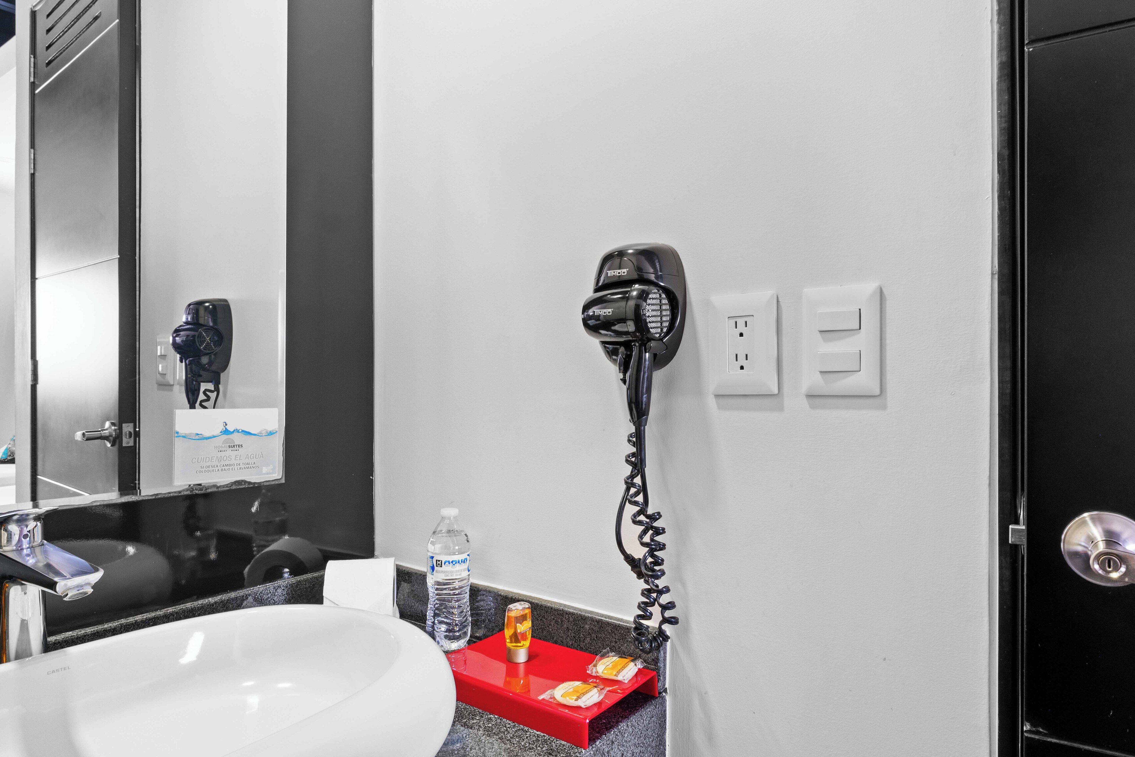 A bathroom with a white sink, a red towel rack, a hairdryer, a bottle of water, and a mirror.