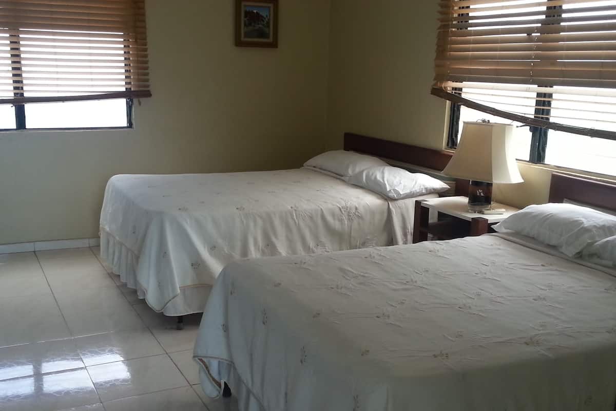 A hotel room with two beds, a nightstand, a lamp, and a window with blinds.