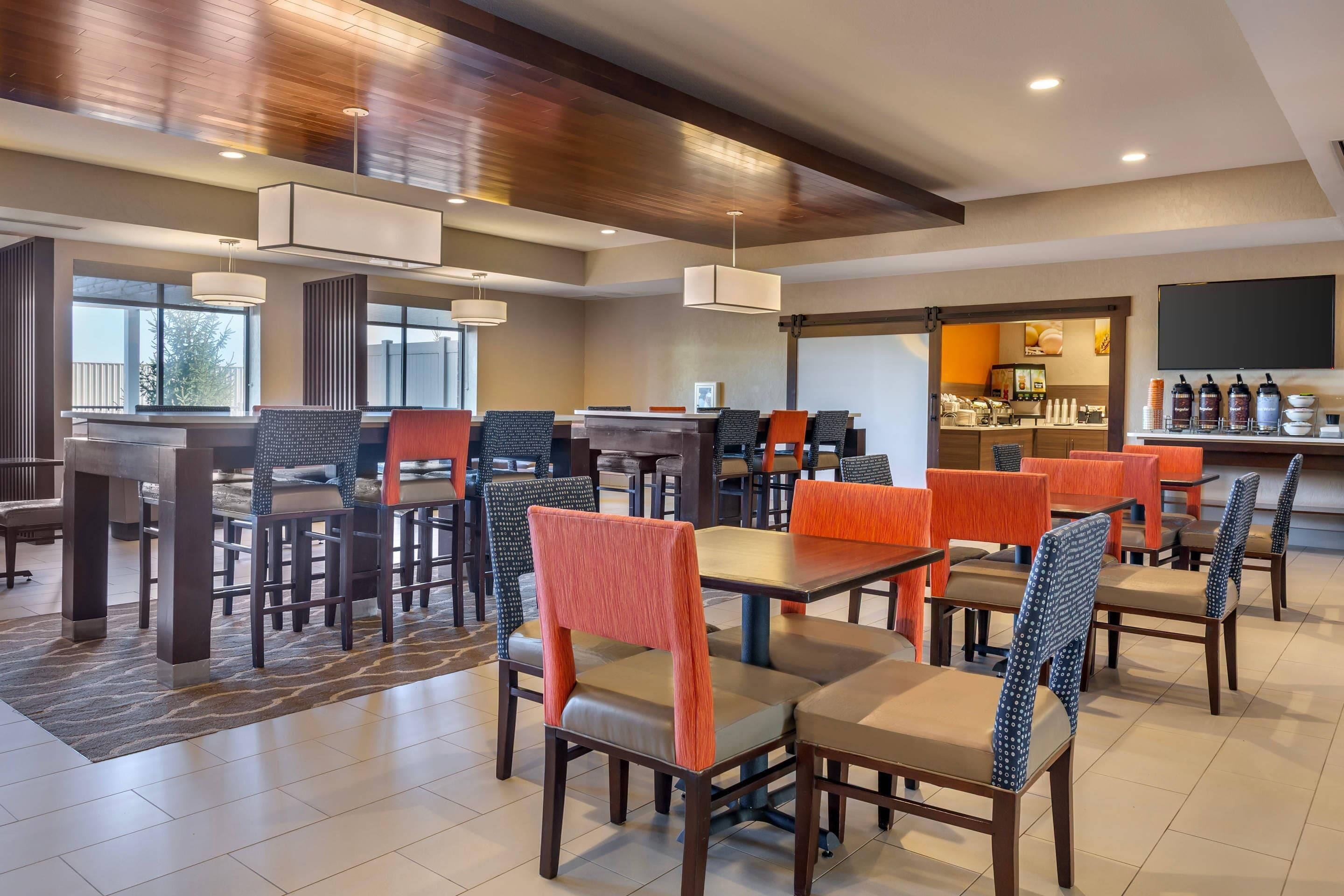 A modern dining area with multiple tables and chairs, a kitchen in the background, and a large screen on the wall.