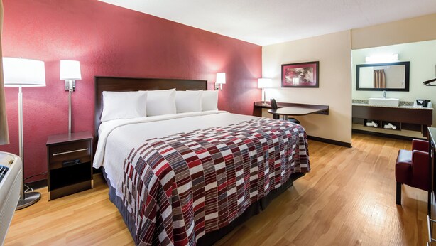A hotel room with a bed, bedside lamps, a desk, a chair, a mirror, and a bathroom vanity. - Red Roof Inn Marion, IN (Marion)
