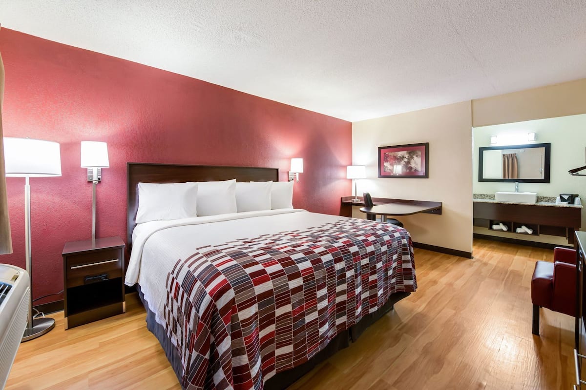 A hotel room with a bed, bedside tables, a desk, a chair, a mirror, and a bathroom with a sink and towel rack.