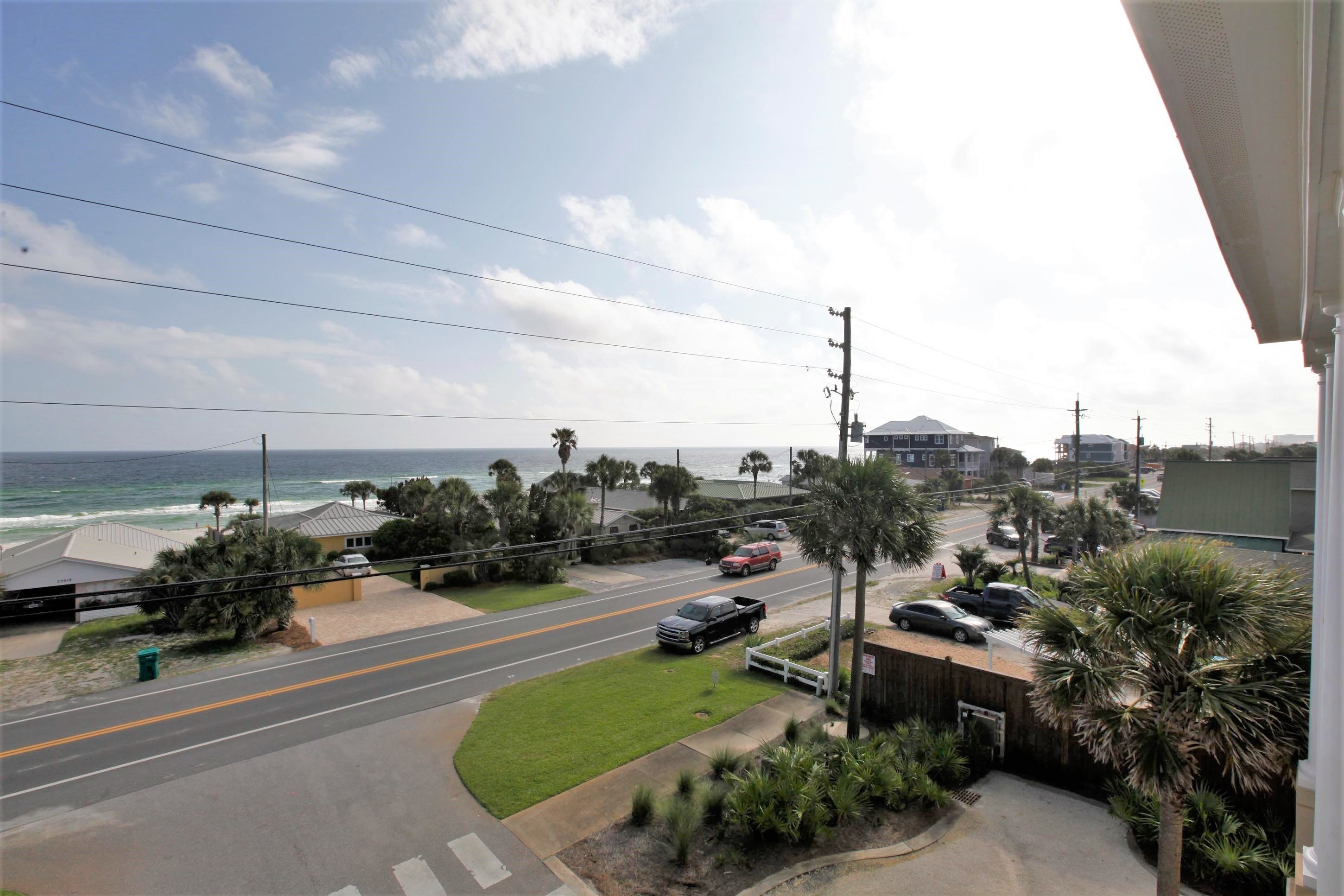 A coastal road with a view of the ocean, palm trees, and a building with a sloped roof.
