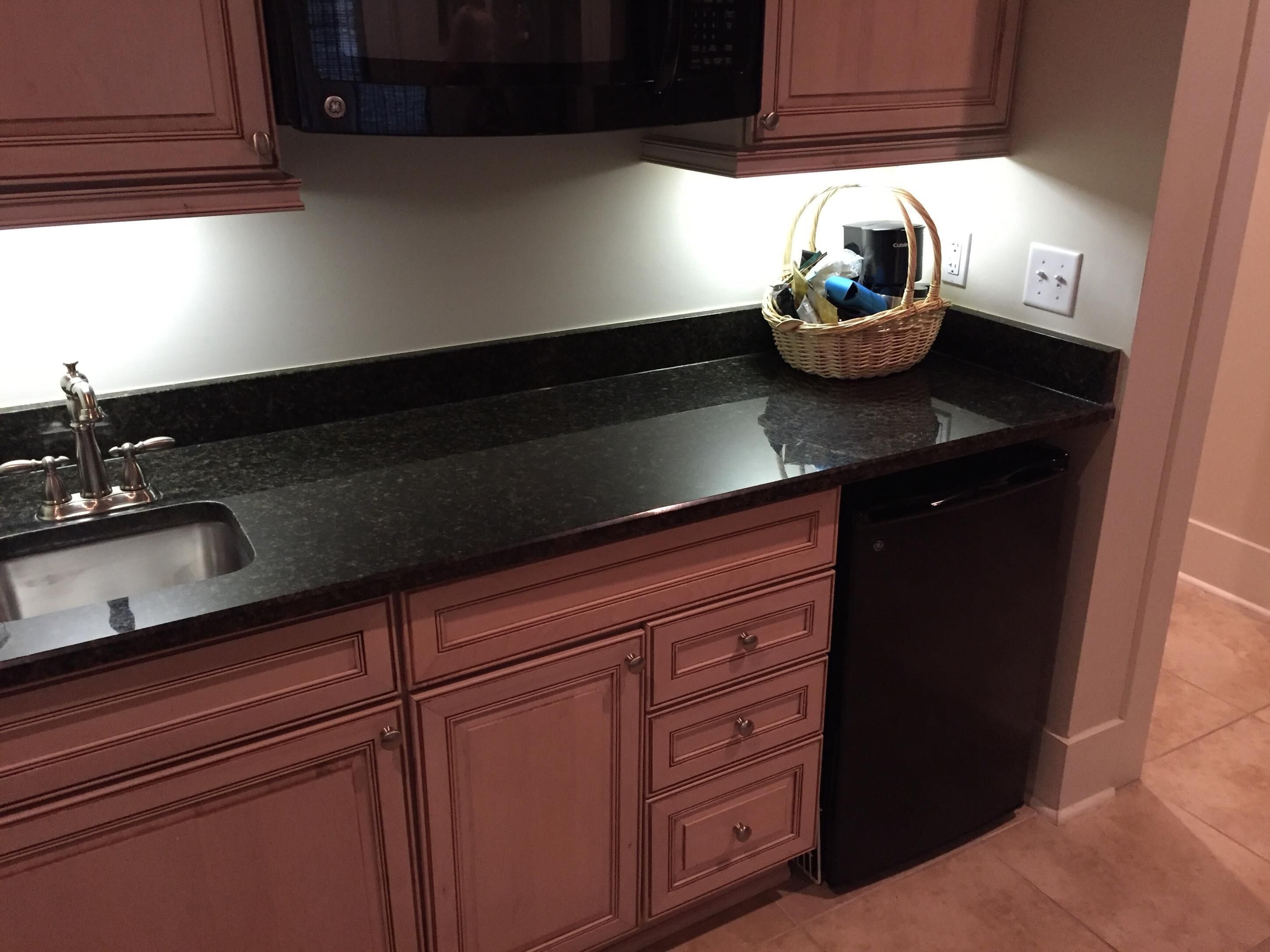 A kitchen with a black countertop, a stainless steel sink, wooden cabinets, a microwave, and a basket on the counter.