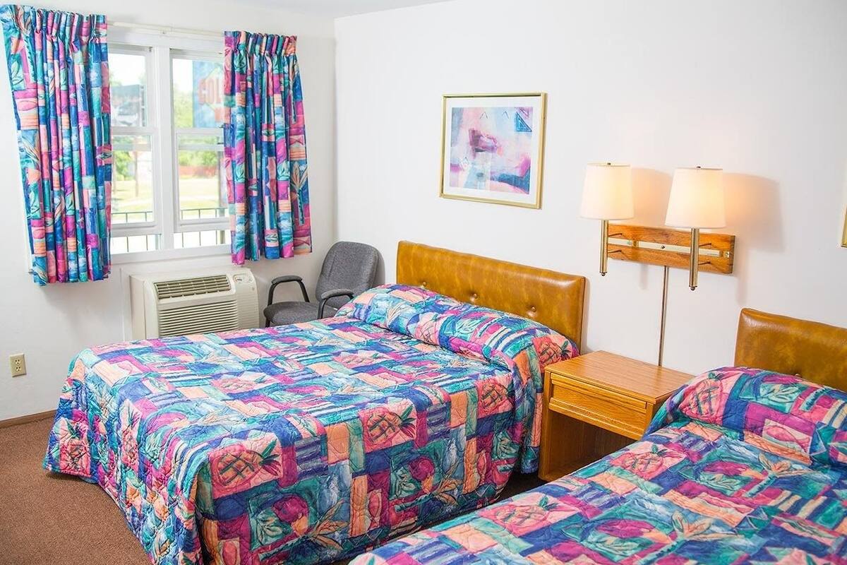 A hotel room with two beds, a desk, a chair, a window with curtains, an air conditioner, and a wall-mounted lamp.