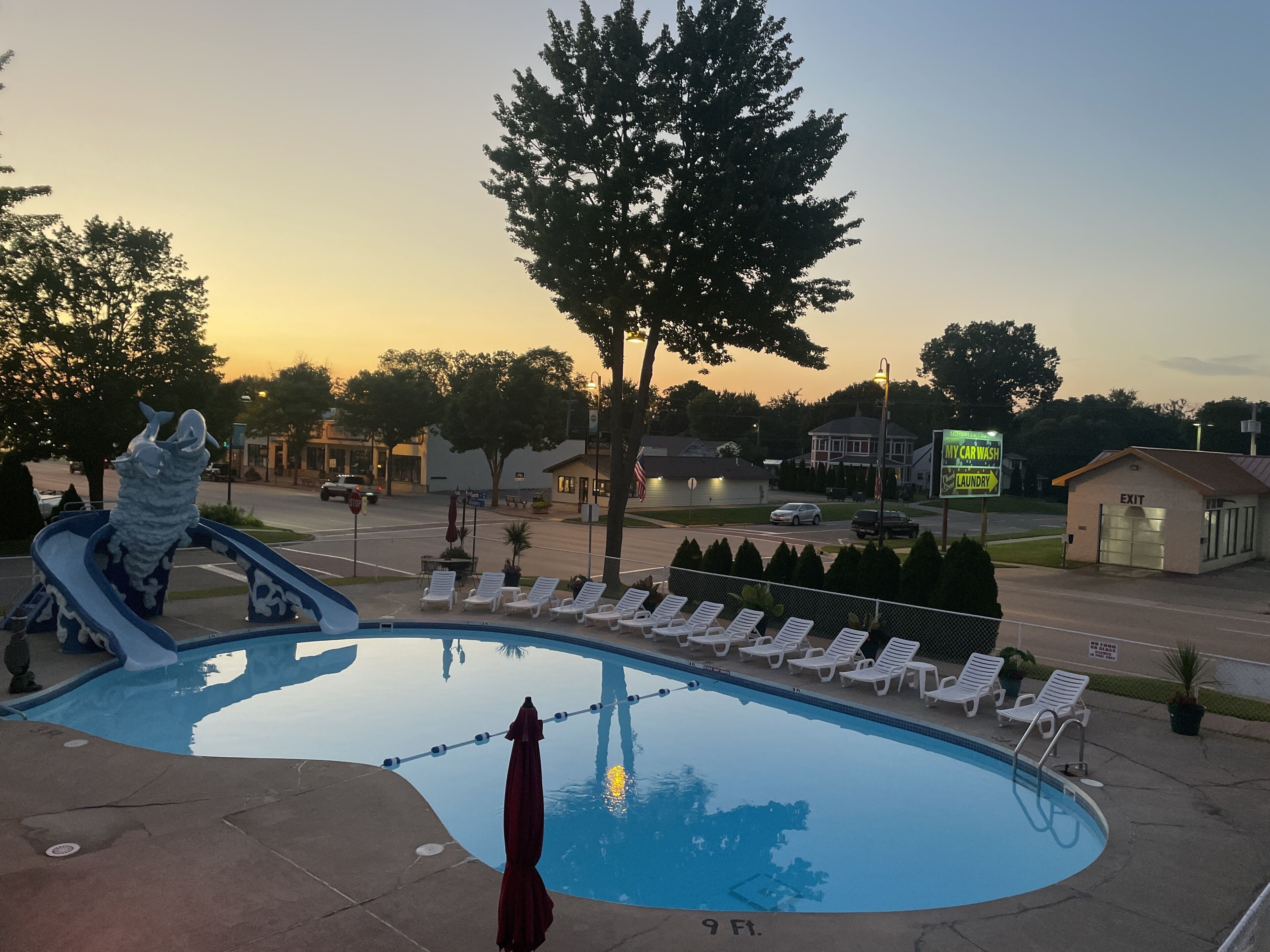 Una piscina con sillones de descanso, una estatua y un letrero de lavado de autos al fondo.