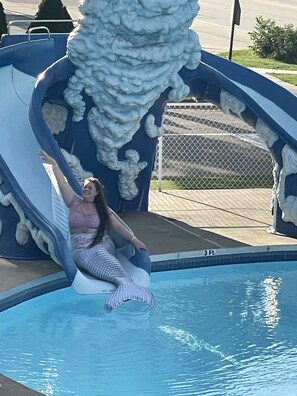 A person in a mermaid costume sitting on a slide at the edge of a pool.