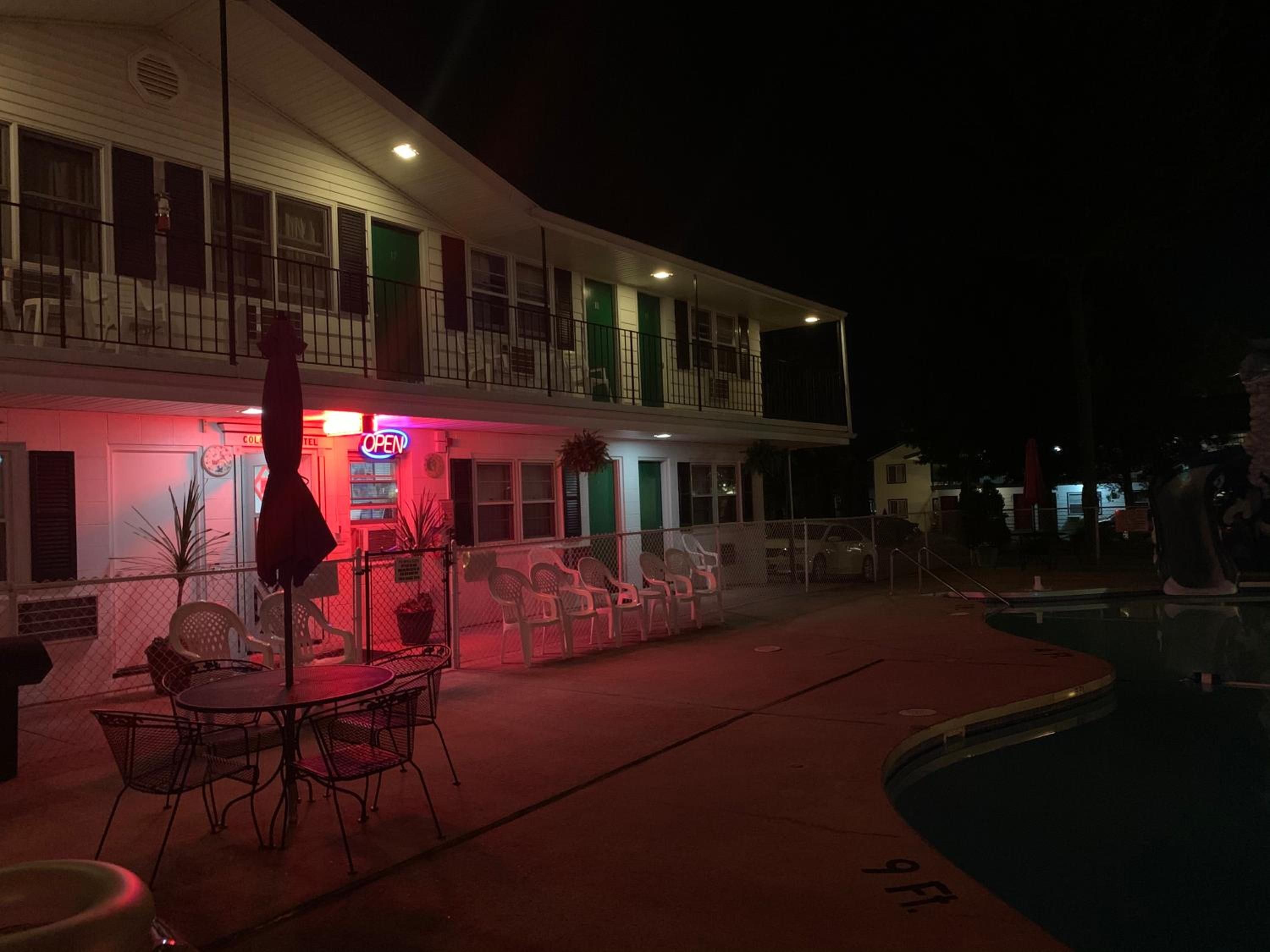 Hotel com piscina, área de estar externa e letreiro de neon indicando que está aberto.