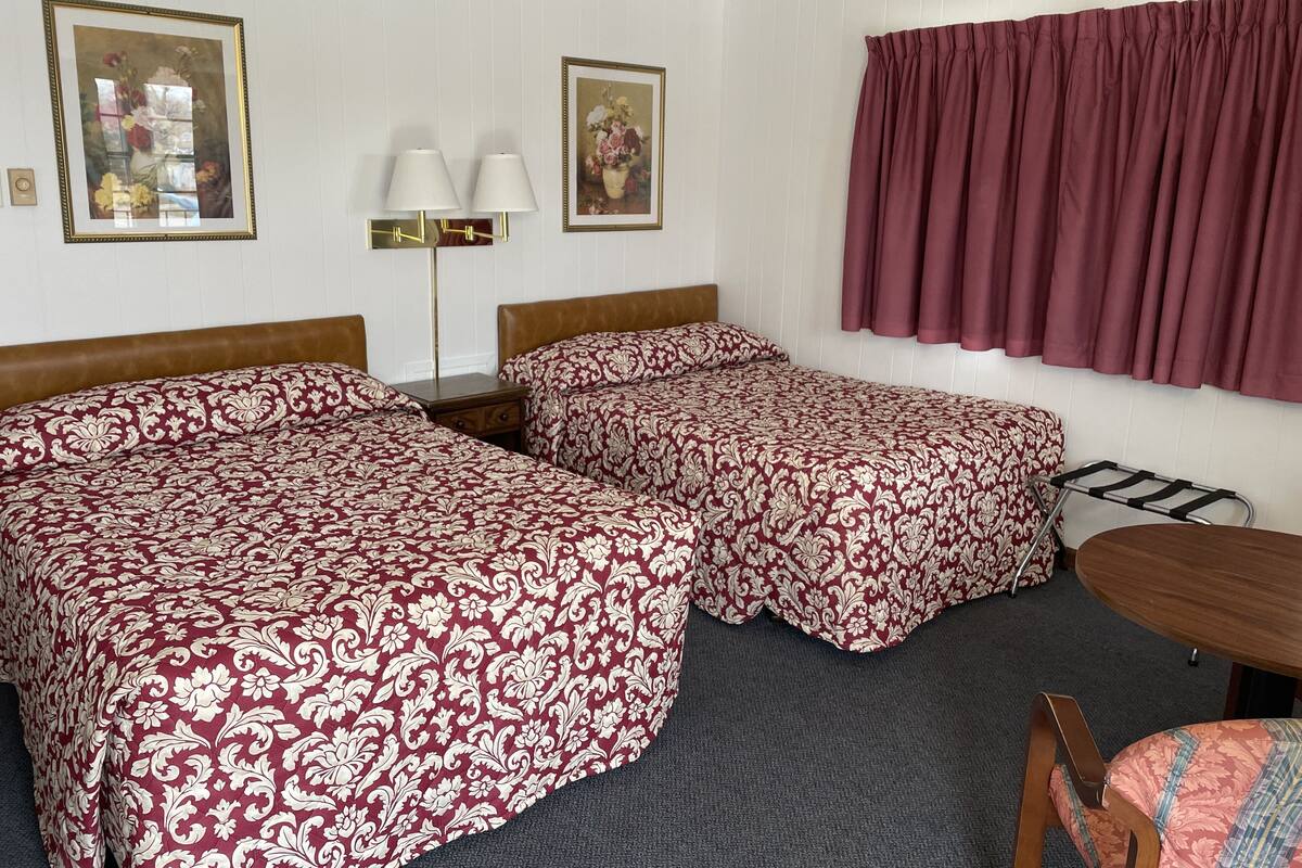 A hotel room with two beds, a desk, and a chair. There are two lamps on the desk and a window with curtains.