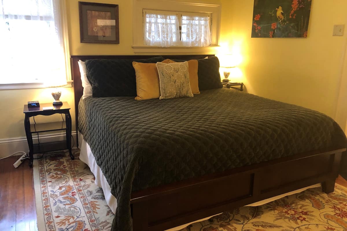A bedroom with a bed, a nightstand, a lamp, a window with lace curtains, and a painting on the wall.