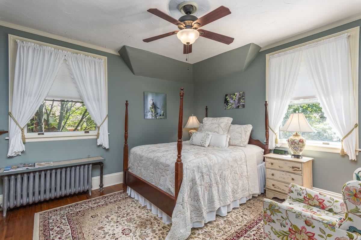 A bedroom with a four-poster bed, a radiator, a wooden nightstand, a chair, and a ceiling fan.