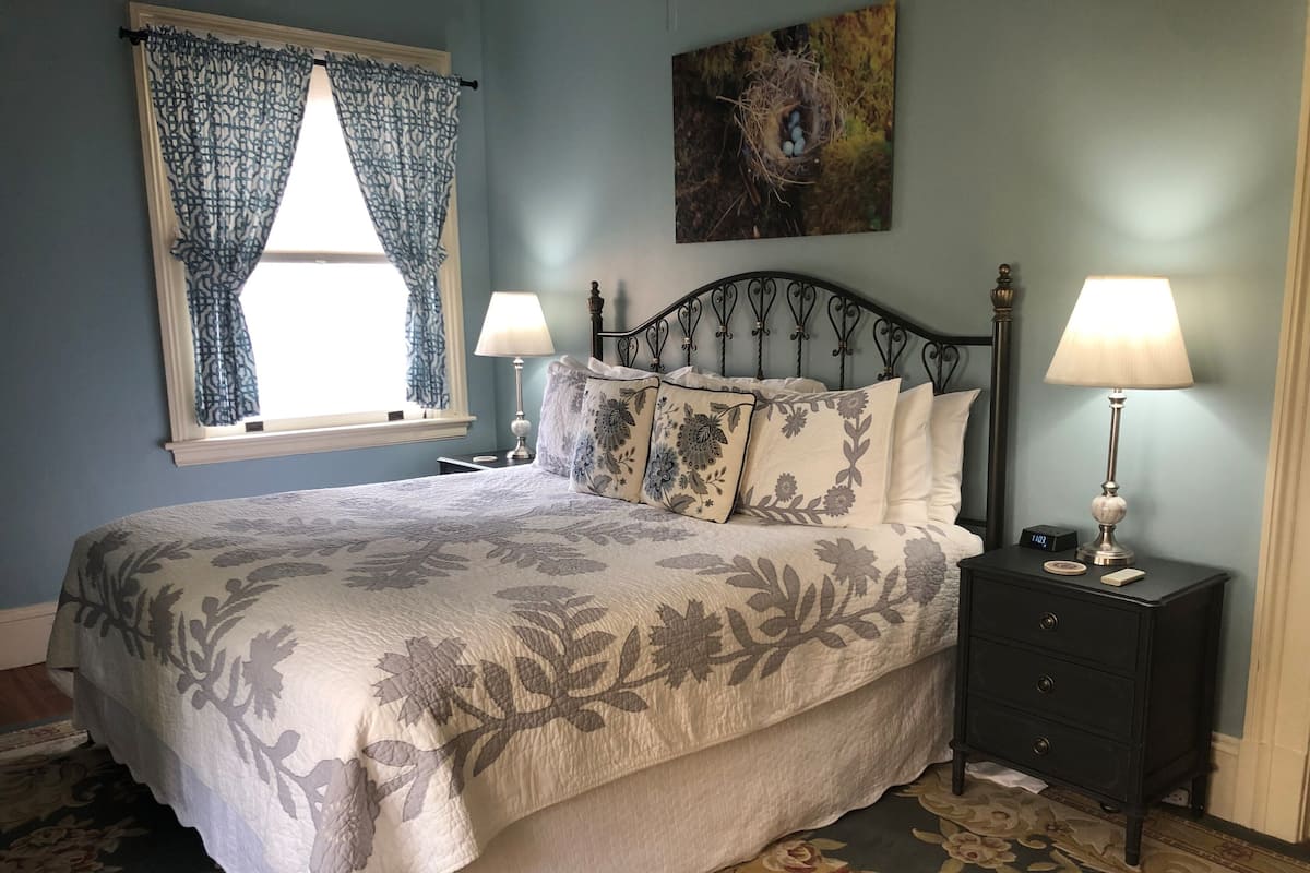 A bedroom with a bed, two bedside tables with lamps, a window with curtains, and a wall-mounted painting.