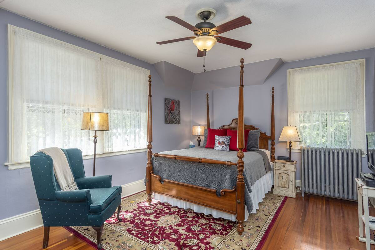 A four-poster bed with a wooden frame and a grey bedspread, a blue armchair with a throw blanket, and a red and white patterned rug.