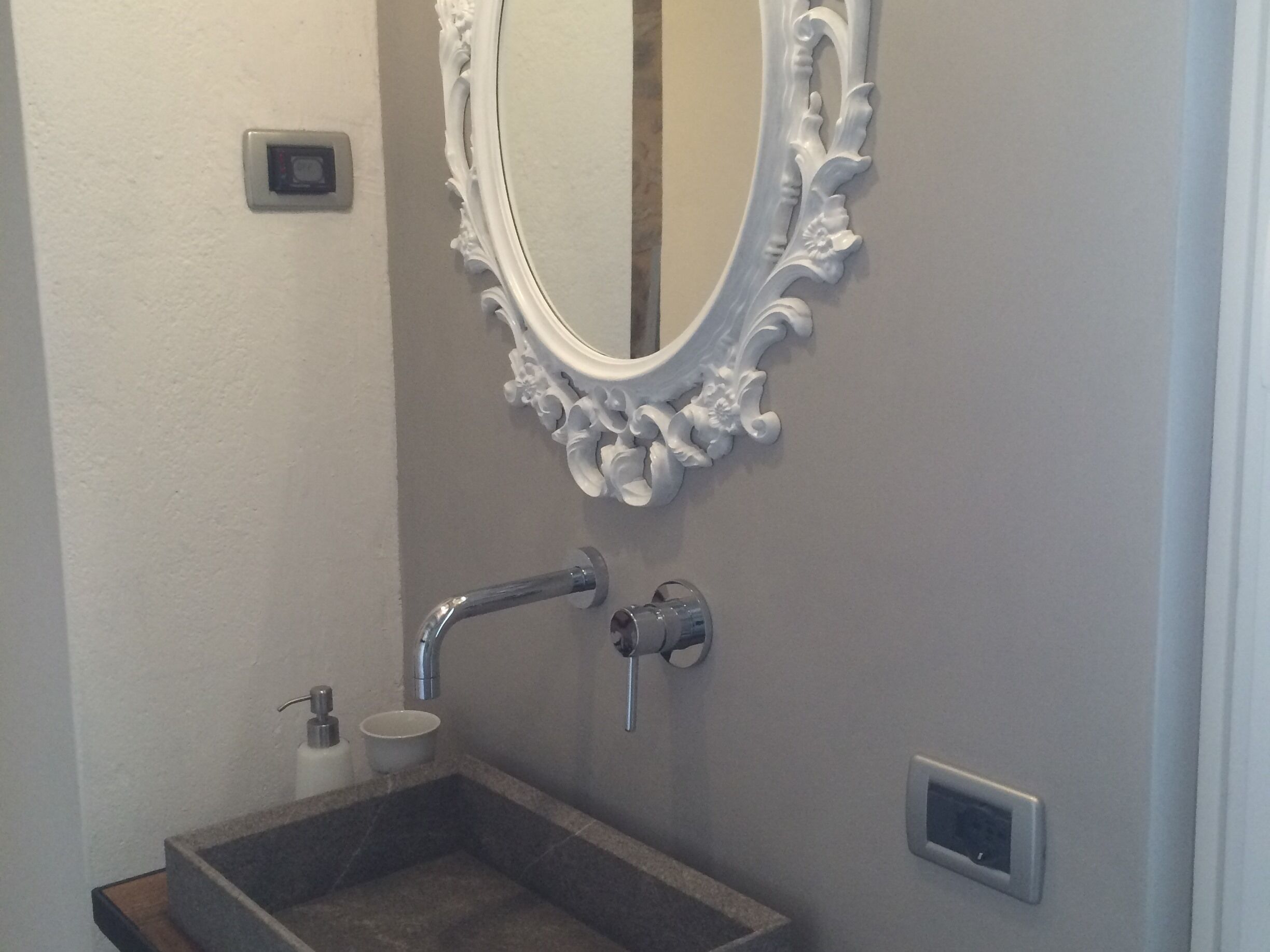 Un bagno con un lavandino rettangolare, uno specchio bianco decorato, un rubinetto a parete e un piccolo dispenser bianco.