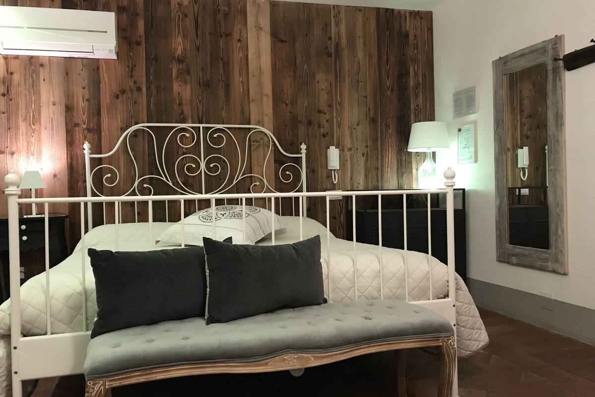 A bedroom with a wooden wall panel, a white metal bed frame, a quilted bedspread, two pillows, a bedside lamp, and a nightstand.