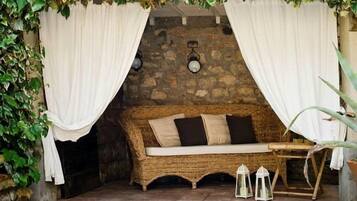 Un espace extérieur accueillant, aménagé avec un canapé en osier, des rideaux blancs, des murs en pierre et des lanternes.