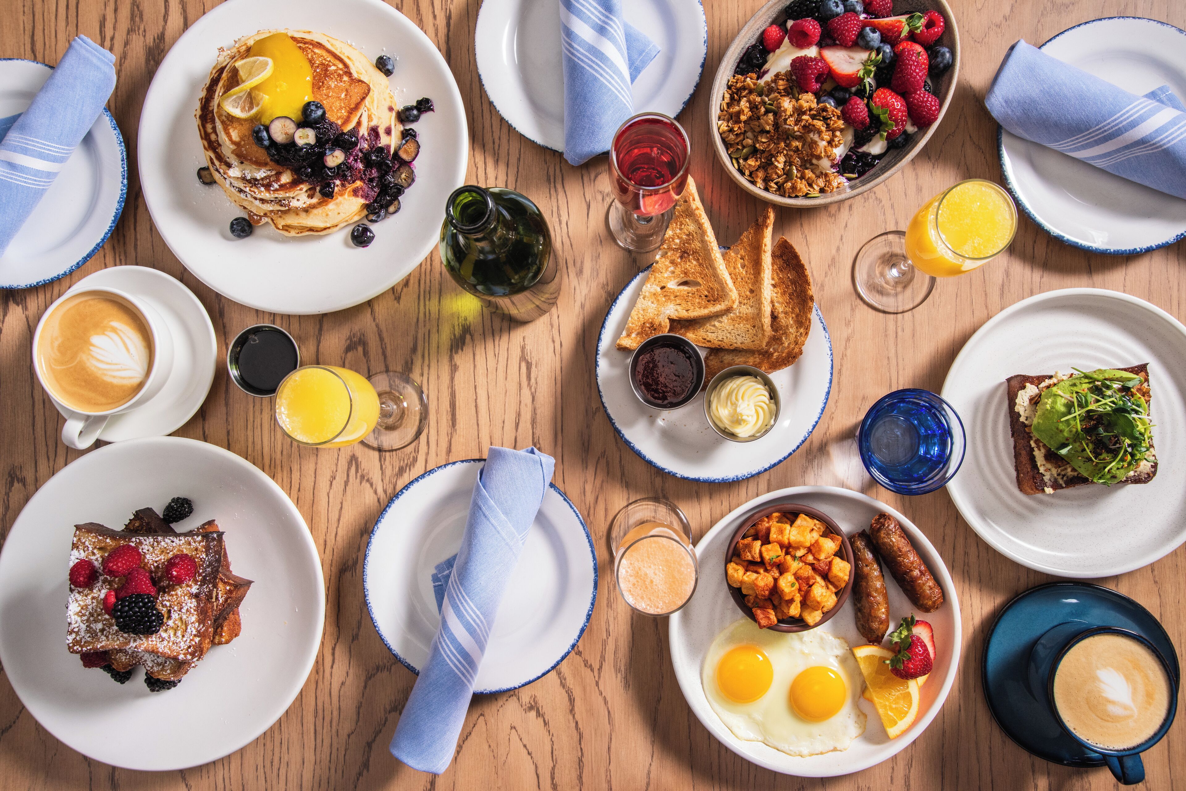 A breakfast setting with pancakes, toast, eggs, sausages, and various fruits and beverages on a wooden table.