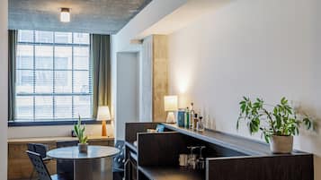 Una habitación de hotel moderna con una mesa redonda, una planta en maceta, un sofá y un amplio ventanal con persianas.
