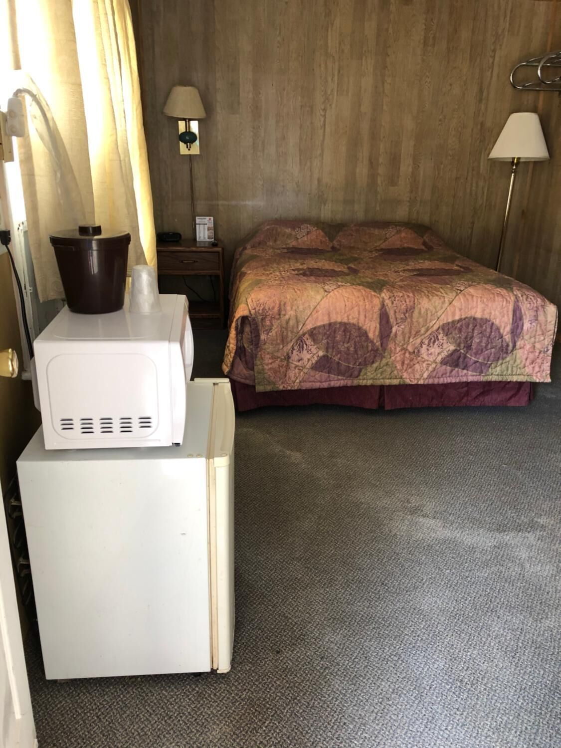 Una habitación de hotel con cama, mesita de noche, lámpara, microondas y cubo de basura.