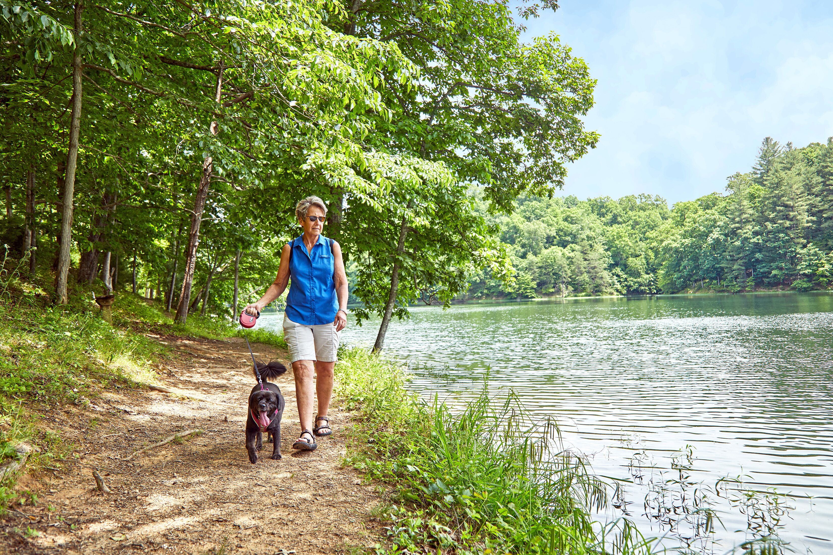 A woman walking a dog on a dirt path beside a lake, surrounded by trees.