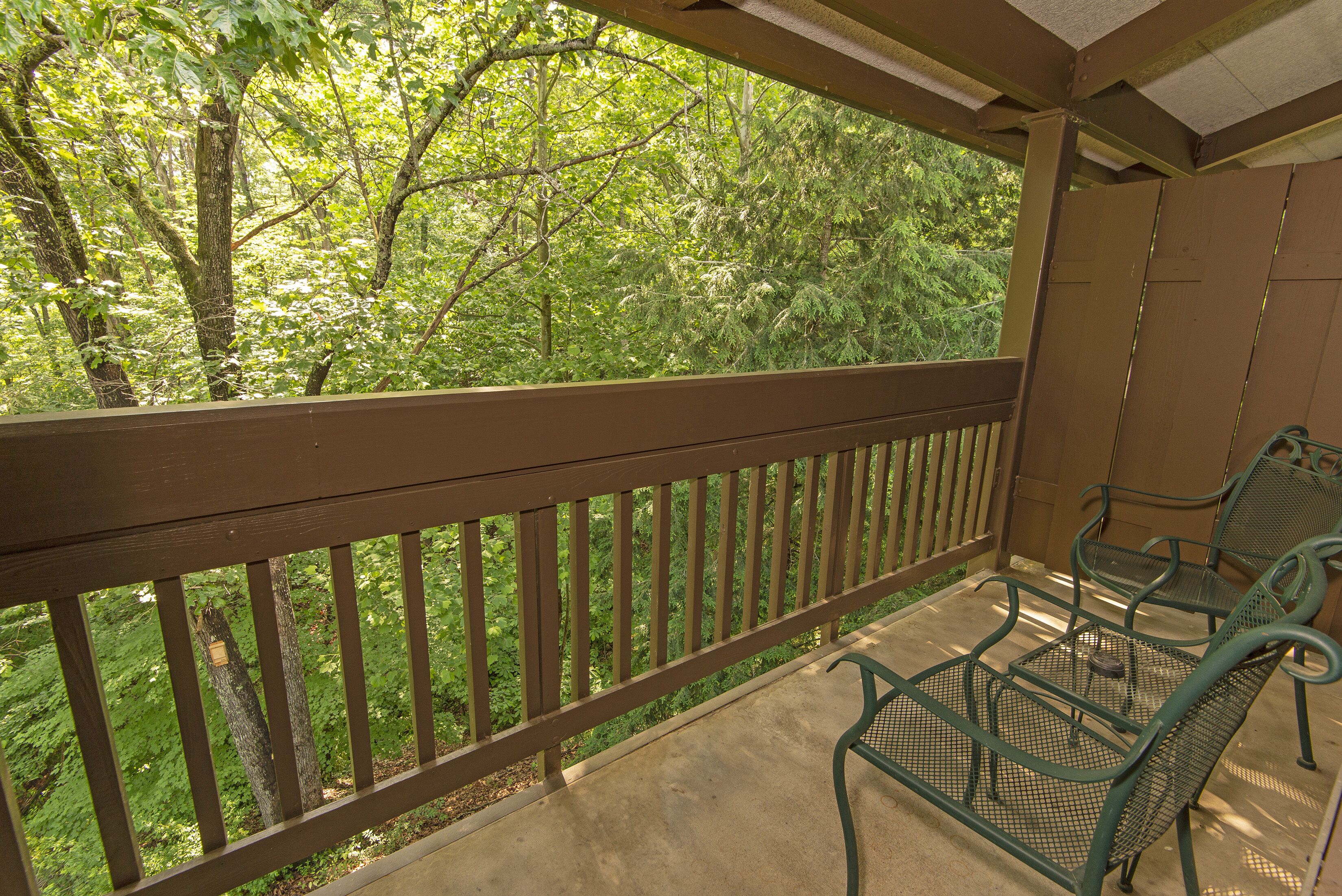 A balcony with a wooden railing, two green metal chairs, and a view of dense green foliage.