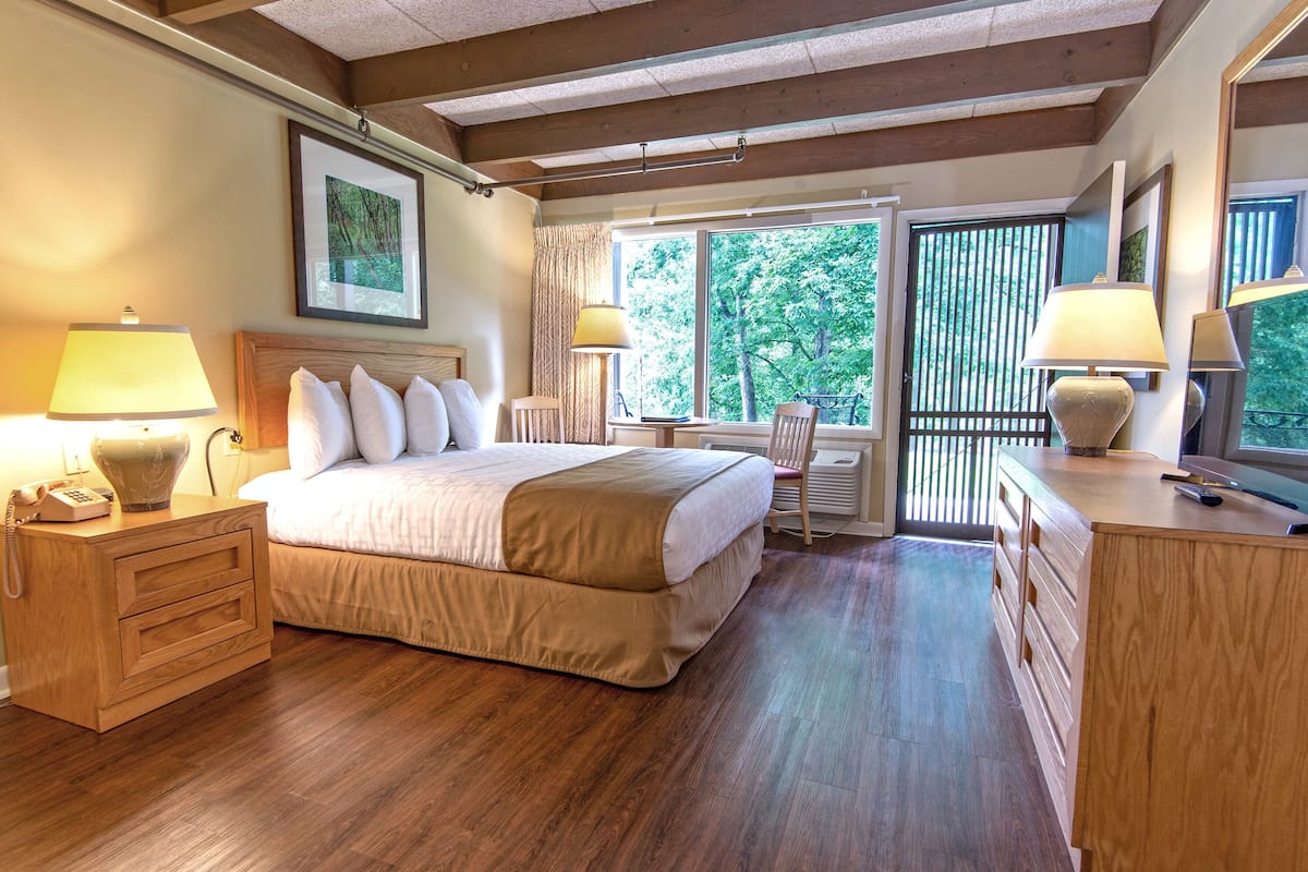 Une chambre avec un grand lit, des lampes de chevet, un bureau et une vue sur les arbres par la fenêtre.