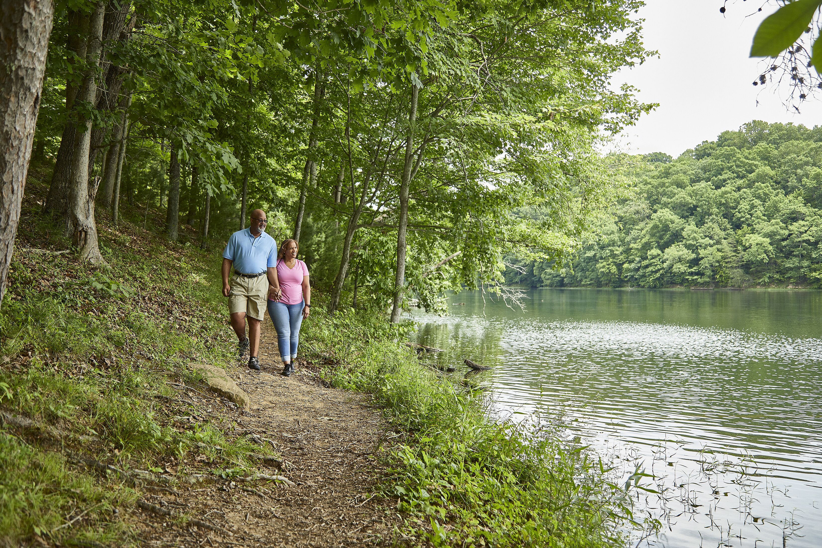 Un couple se promène sur un sentier de terre bordant un lac, entouré d’arbres.