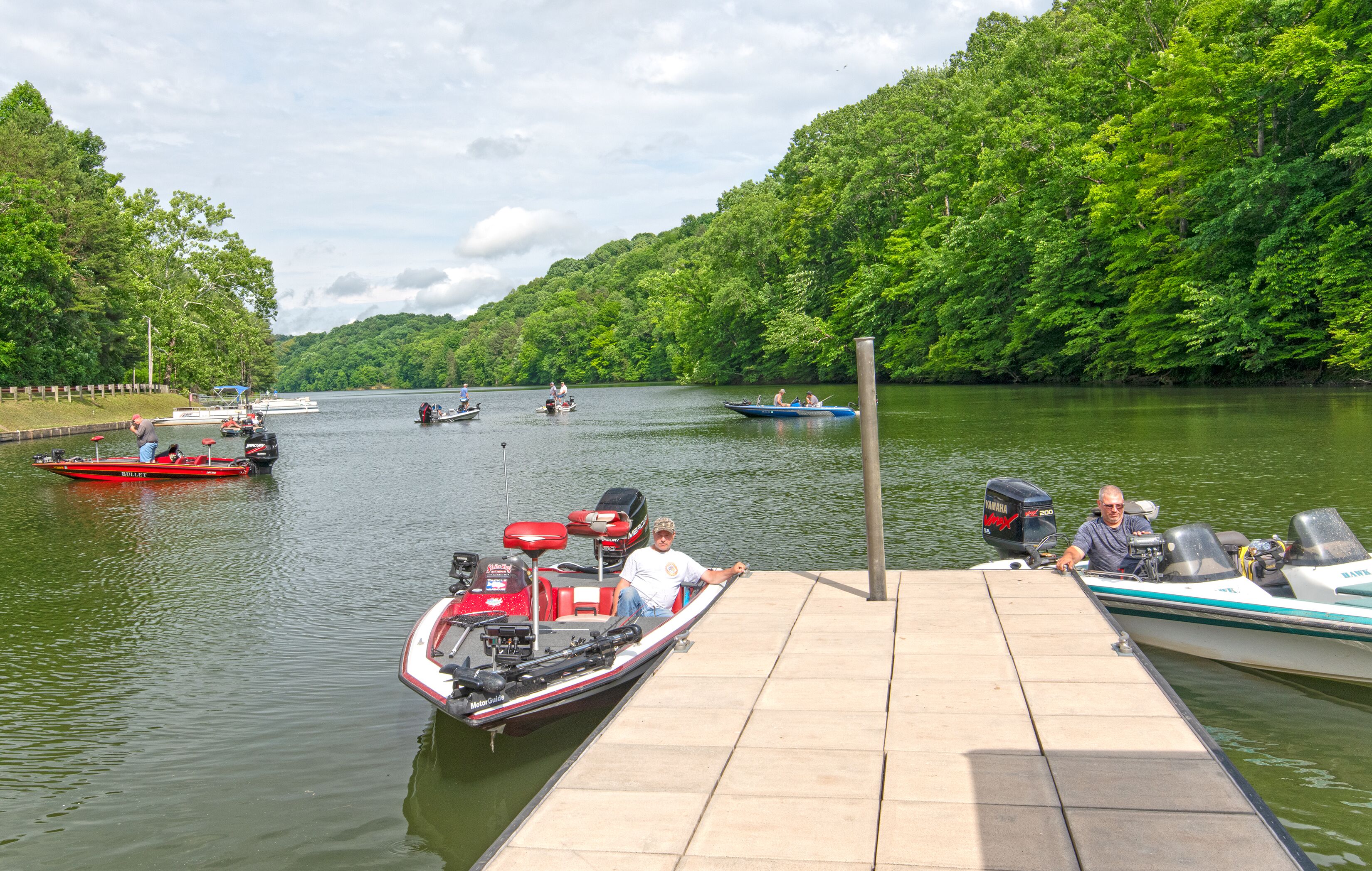 A dock with several boats, including a red and a white one, and a forested hillside in the background.