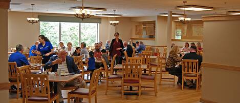 A spacious dining area with wooden tables and chairs, large windows allowing natural light, and a high ceiling with hanging lights.