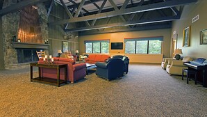 Un vaste salon doté d’une cheminée en pierre, de sièges confortables, d’un piano et de grandes fenêtres offrant une vue sur l’extérieur.