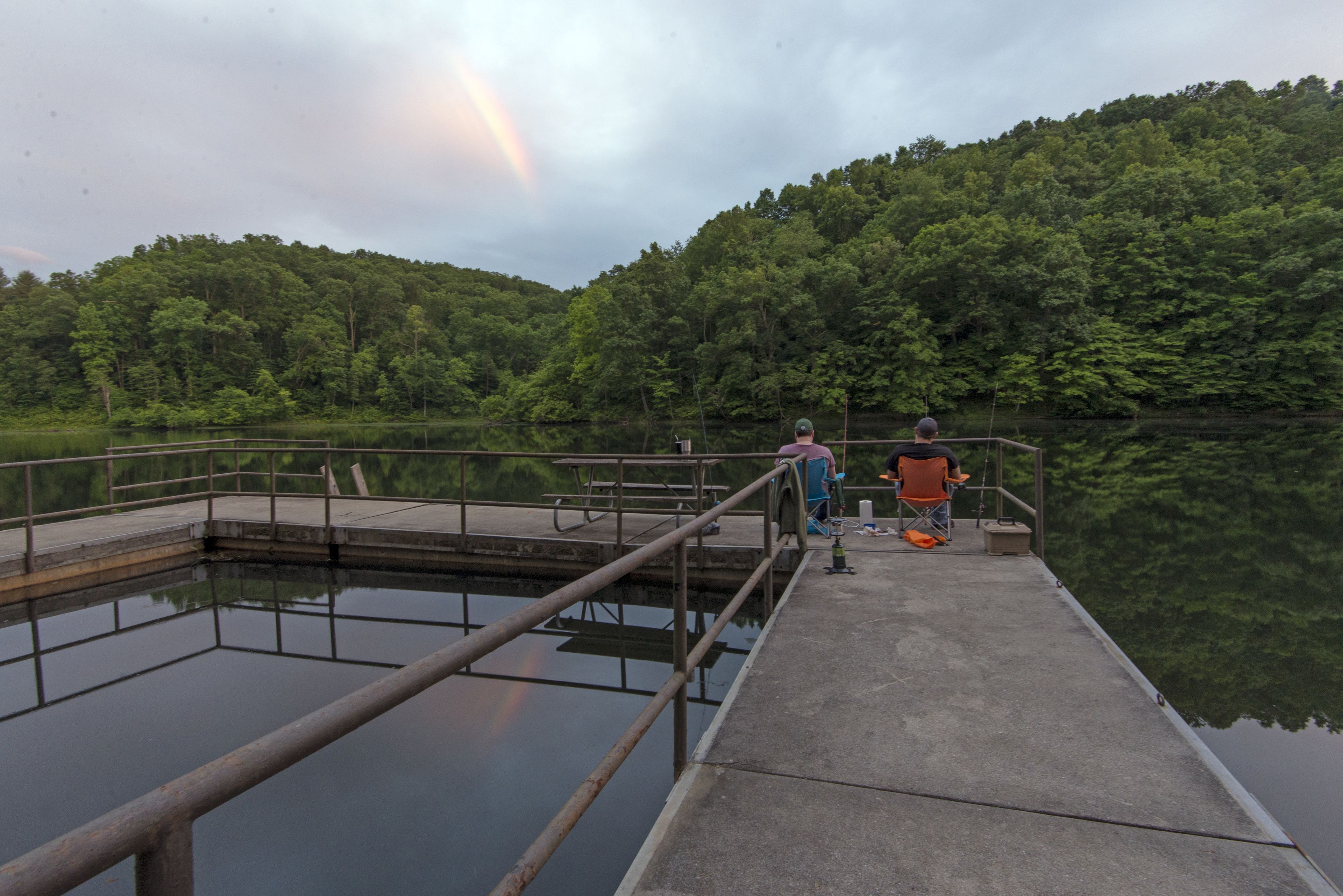 A concrete pier extending into a calm lake with a forested hill in the background.