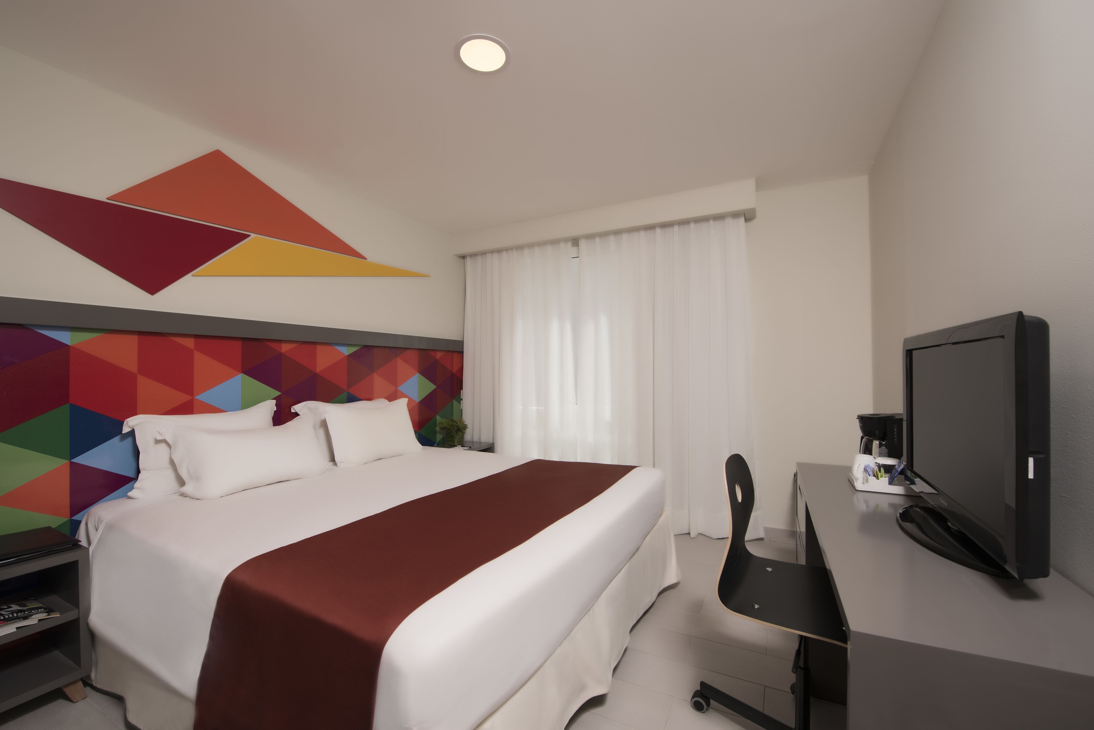 A hotel room with a bed, a desk with a television, and a colorful geometric wall design.