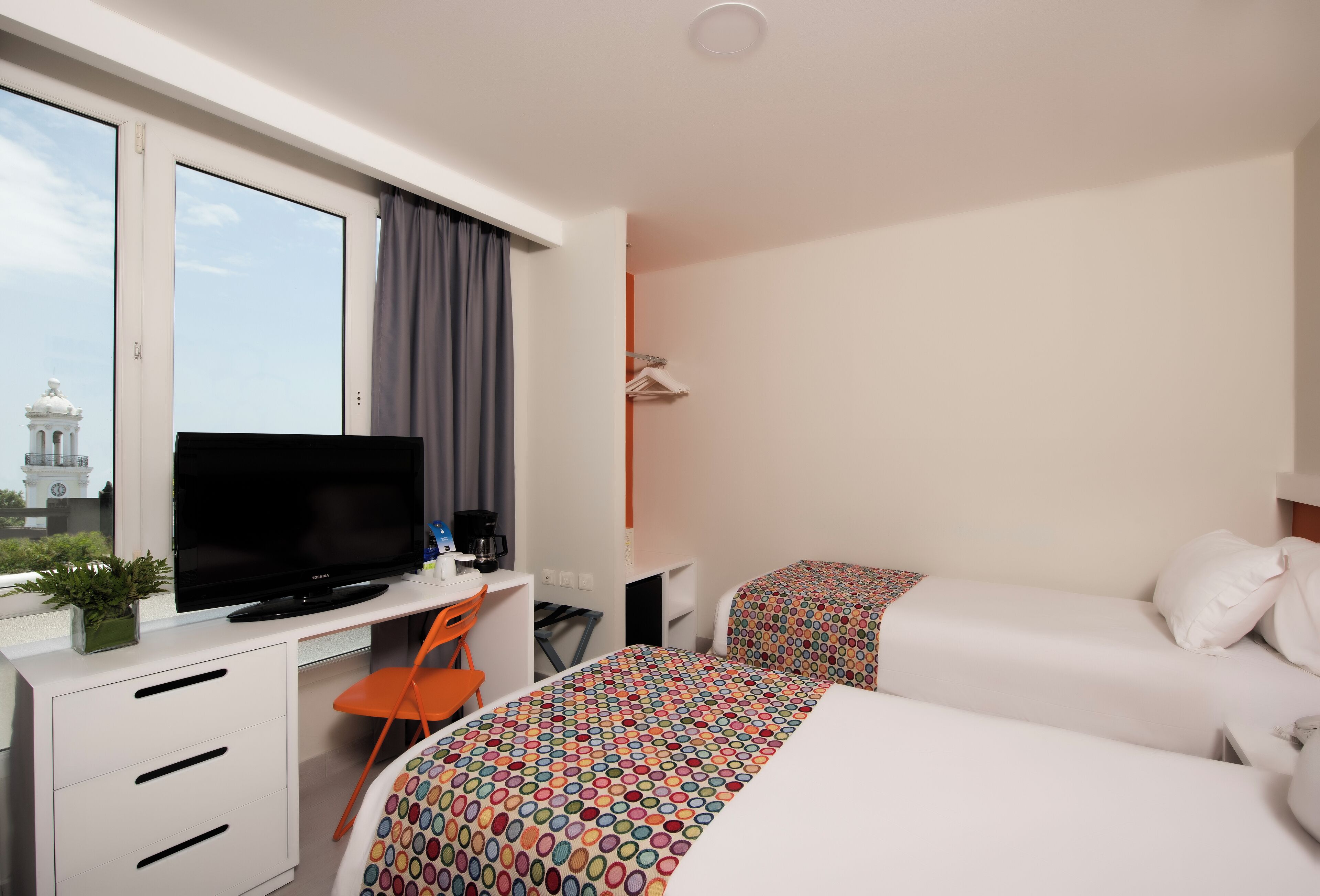 A hotel room with a bed, a desk with a television, a chair, and a window with curtains.
