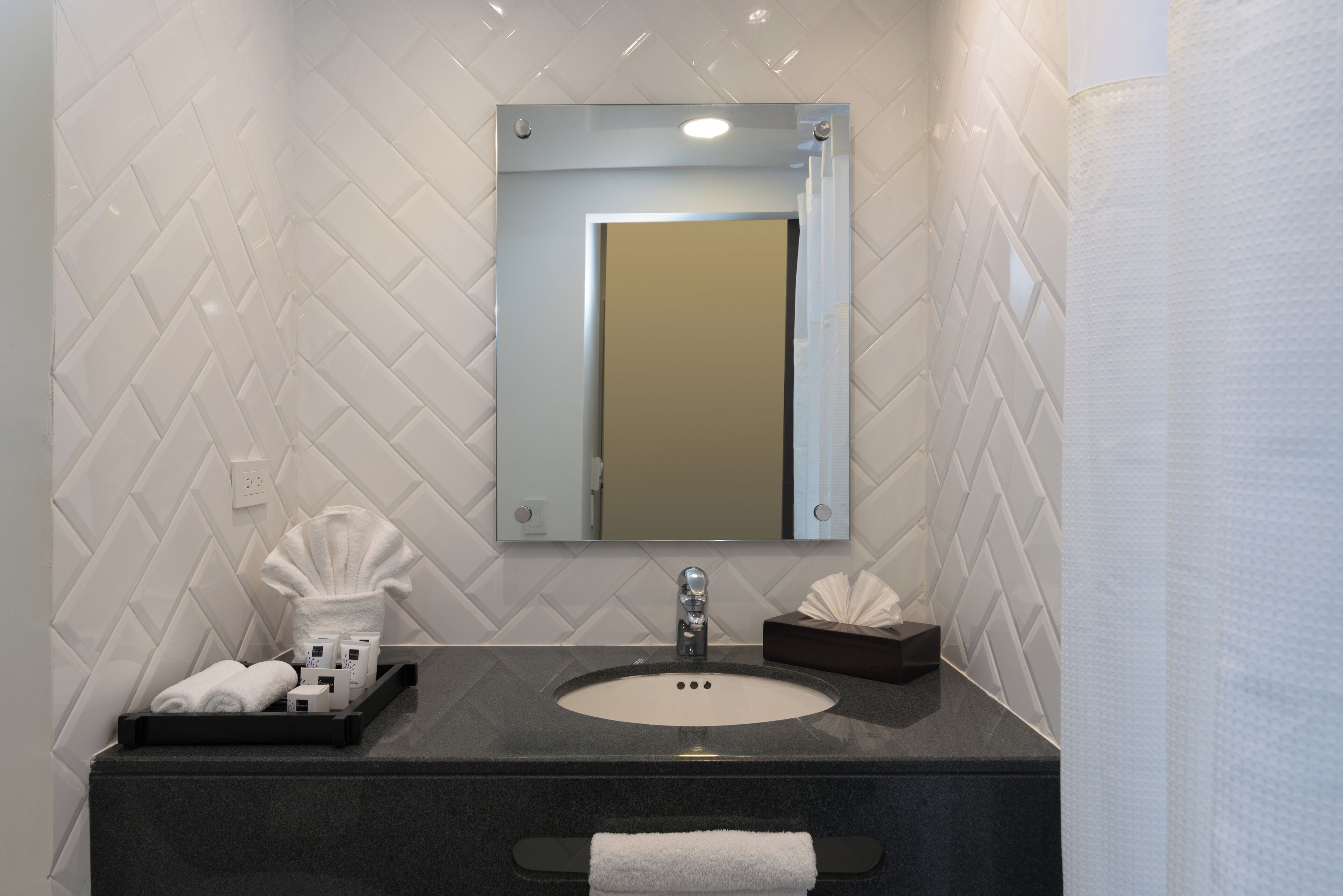 Une salle de bain avec un mur recouvert de carreaux blancs disposés en motif chevron, un plan de toilette noir, un lavabo rond, un miroir et un porte-serviettes.