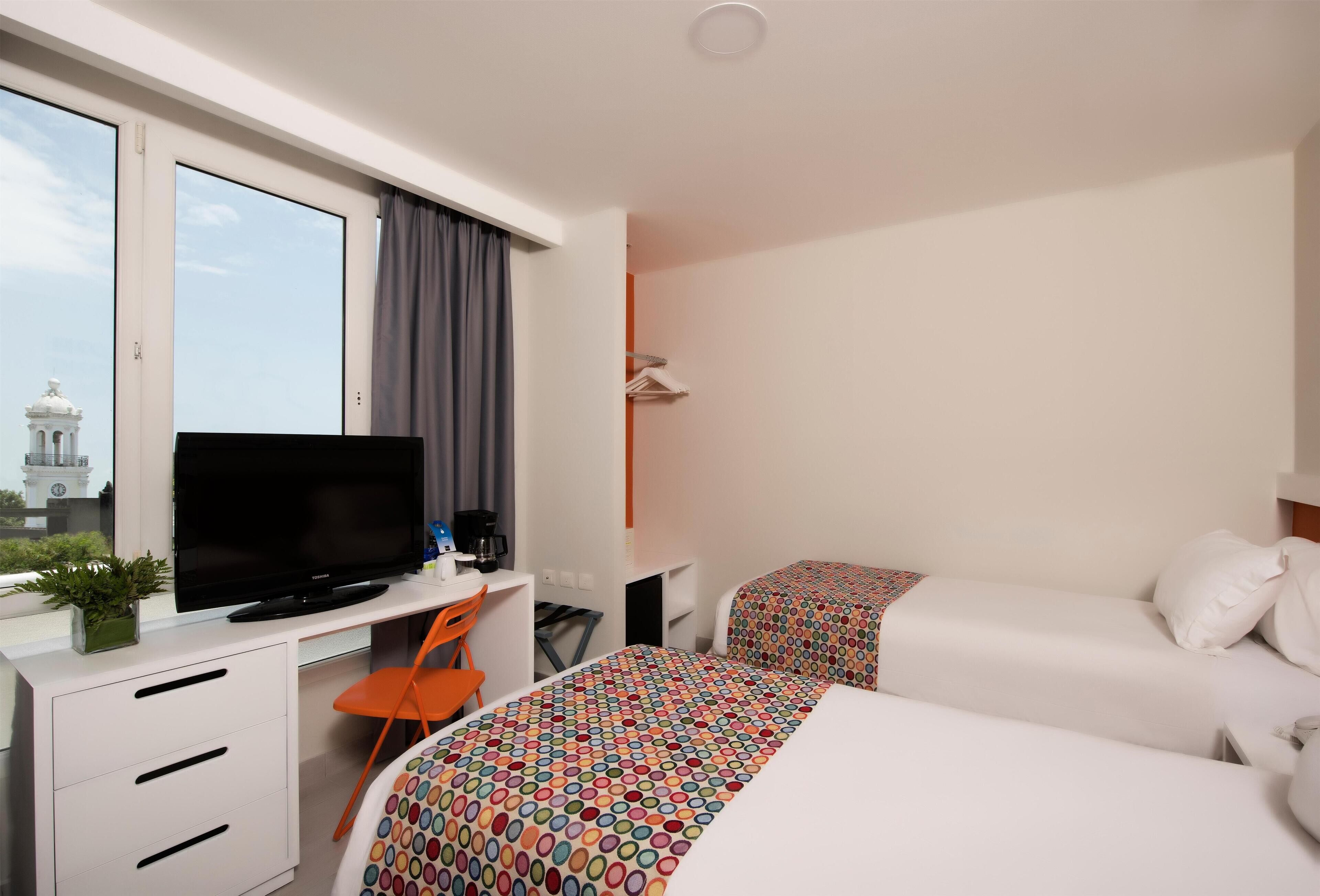 Habitación de hotel con un ventanal grande, un escritorio con televisor de pantalla plana, una silla, dos camas con ropa de cama colorida y una planta pequeña en el escritorio.