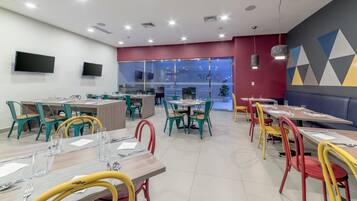 A modern restaurant with multiple dining tables and chairs, a large TV screen, and a glass wall offering a view of the interior.