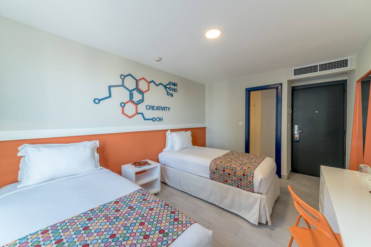 Ein Hotelzimmer mit zwei Betten, einem Schreibtisch und einem Stuhl. An der Wand befindet sich eine Dekoration mit einer Molekülstruktur und dem Wort 'CREATIVITY'.