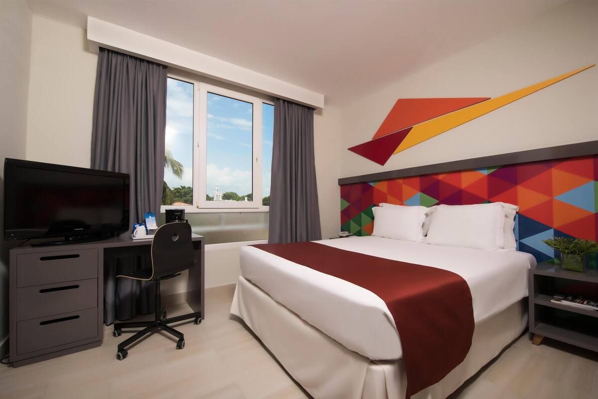 Ein Hotelzimmer mit Bett, Schreibtisch mit Fernseher, Stuhl, Fenster mit Vorhängen und farbigem Wanddesign.
