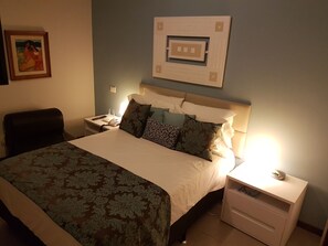 A hotel room with a large bed, a nightstand with a lamp, a chair, and a wall-mounted artwork.
