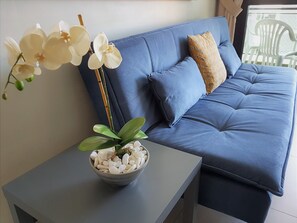 Um sofá azul com uma almofada estampada e uma orquídea branca numa tigela sobre uma mesinha.