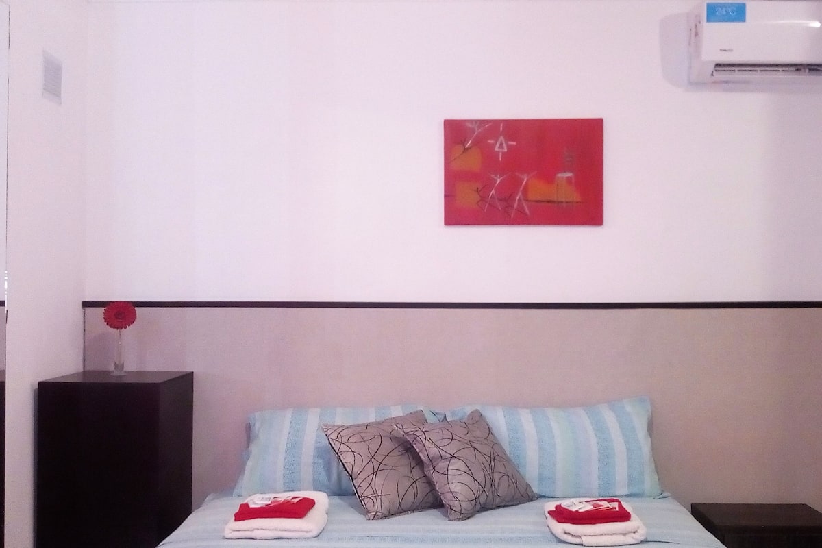 A hotel room with a bed, a nightstand, a red artwork on the wall, and an air conditioner.